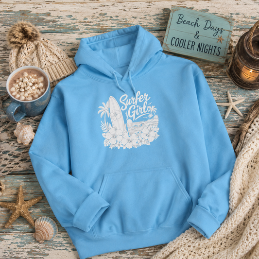 A light blue pullover hoodie with a white 'Surfer Girl' graphic featuring a surfboard, palm tree, and flowers, styled in a beach-themed flat lay on a rustic wooden surface with a knit hat, mug, lantern, and starfish.