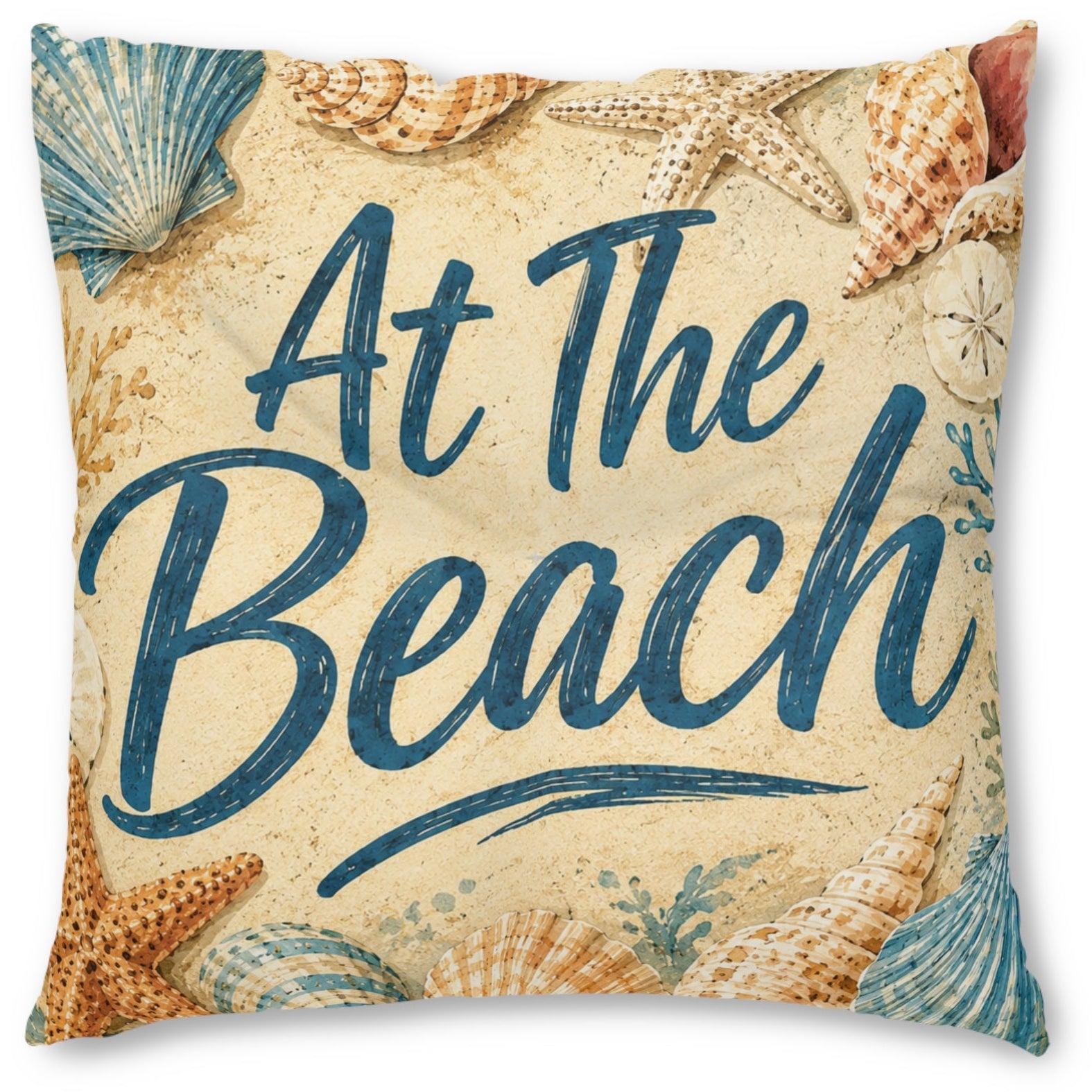 A square decorative throw pillow featuring a beach-themed print with the words 'At The Beach' in dark blue script, surrounded by seashells and starfish on a sandy beige background.