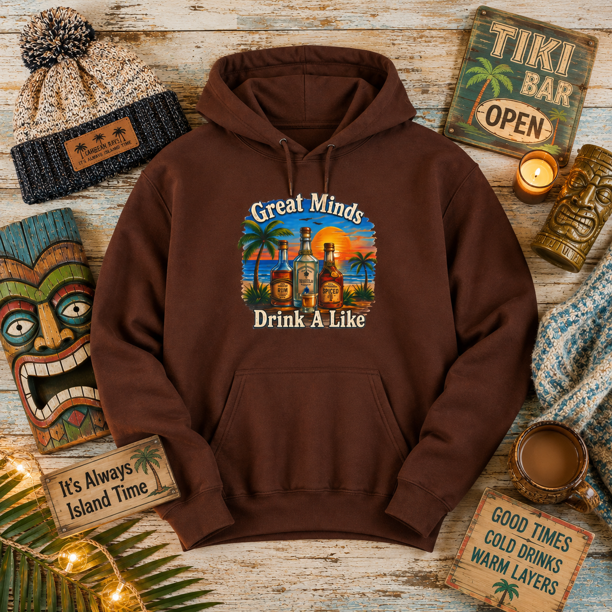 A brown pullover hoodie laid flat on a rustic wood surface, surrounded by tropical-themed props including a beanie, tiki sign, mask, candle, mug, and small signs. The hoodie has a colorful front graphic with the text 'Great Minds Drink A Like' and an illustration of liquor bottles on a beach at sunset.