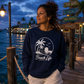 A woman wearing a navy blue crewneck sweatshirt with a white 'Beach Life' graphic stands on a wooden dock at dusk by the water, with a signpost in the background reading 'Tiki Bar', 'Beach', 'Boat Rentals', and 'Paradise'.