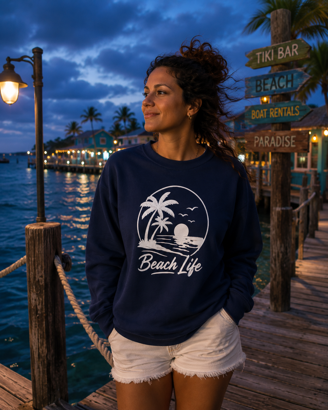 A woman wearing a navy blue crewneck sweatshirt with a white 'Beach Life' graphic stands on a wooden dock at dusk by the water, with a signpost in the background reading 'Tiki Bar', 'Beach', 'Boat Rentals', and 'Paradise'.