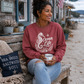 A woman sitting on a wooden bench on a beach street, wearing a dusty rose crewneck sweatshirt with a white 'Ocean Life' seahorse graphic. She is holding a white mug, with a 'Sea Breeze Saltwater Soul' pillow on the bench next to her.
