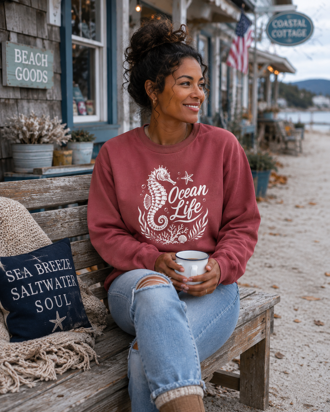 A woman sitting on a wooden bench on a beach street, wearing a dusty rose crewneck sweatshirt with a white 'Ocean Life' seahorse graphic. She is holding a white mug, with a 'Sea Breeze Saltwater Soul' pillow on the bench next to her.