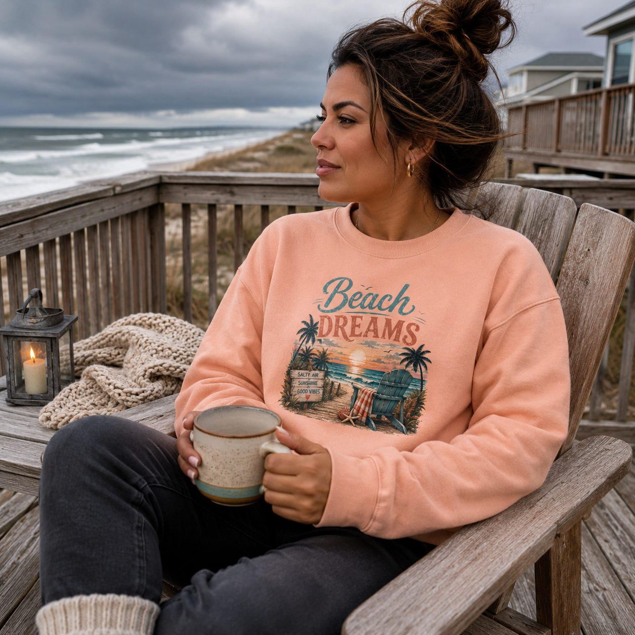 A woman sits in a wooden chair on a beach deck holding a mug, wearing a peach crewneck sweatshirt with a 'Beach Dreams' graphic print. The background shows the ocean, cloudy sky, and a cozy lantern on the table.