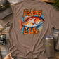 Brown t-shirt with 'Fishing is Life' design, cap, and tumblers on a wooden surface.