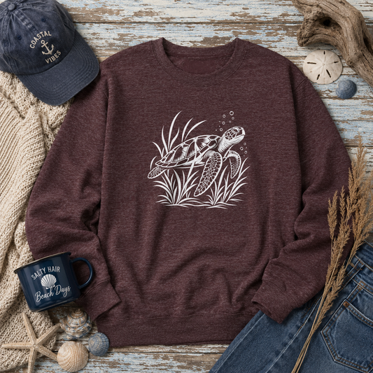 A heathered burgundy crewneck sweatshirt with a white sea turtle graphic, styled in a nautical flat lay with a navy cap, denim shorts, a mug, and beach-themed props on a distressed wood background.