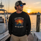 A man wearing a black crewneck sweatshirt with a sunset fishing graphic and the text 'LIFE IS BETTER ON THE WATER' stands on a wooden dock next to a boat at sunset.