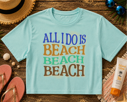 Light blue t-shirt with 'All I Do is Beach' text on a wooden surface with beach accessories.