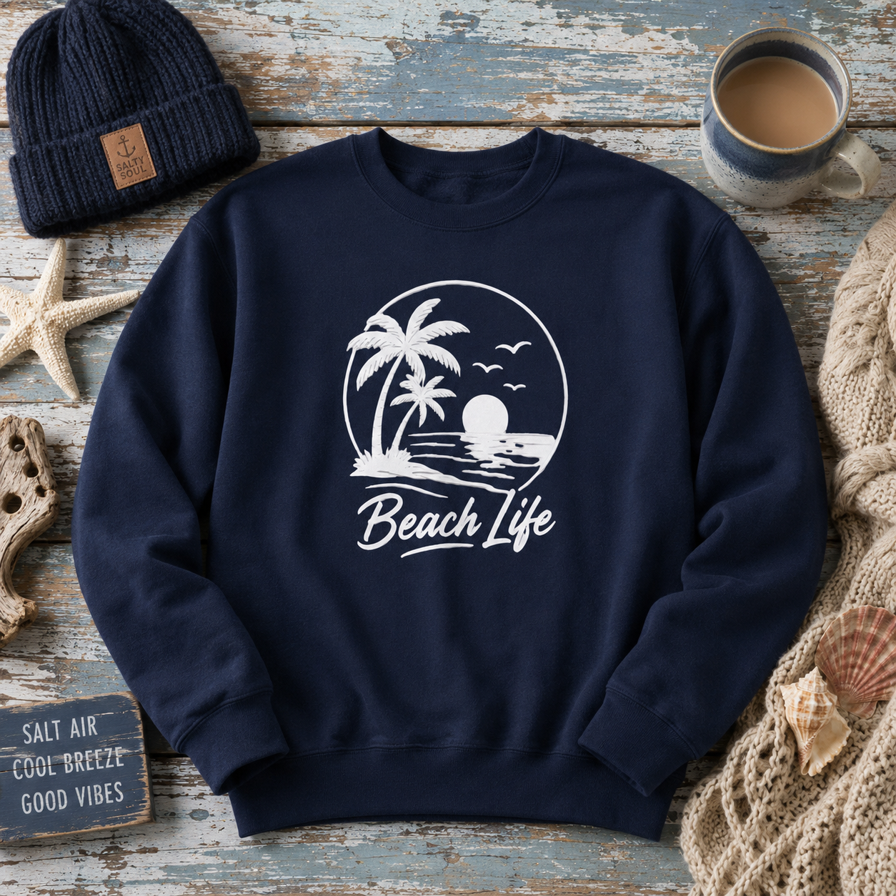 A navy blue crewneck sweatshirt with a white graphic print of a beach scene and the words 'Beach Life'. The sweatshirt is styled in a flat-lay arrangement on a weathered blue wooden surface, surrounded by a navy beanie, a cup of coffee, a starfish, a small sign, and a knitted blanket.