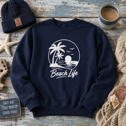 A navy blue crewneck sweatshirt with a white graphic print of a beach scene and the words 'Beach Life'. The sweatshirt is styled in a flat-lay arrangement on a weathered blue wooden surface, surrounded by a navy beanie, a cup of coffee, a starfish, a small sign, and a knitted blanket.