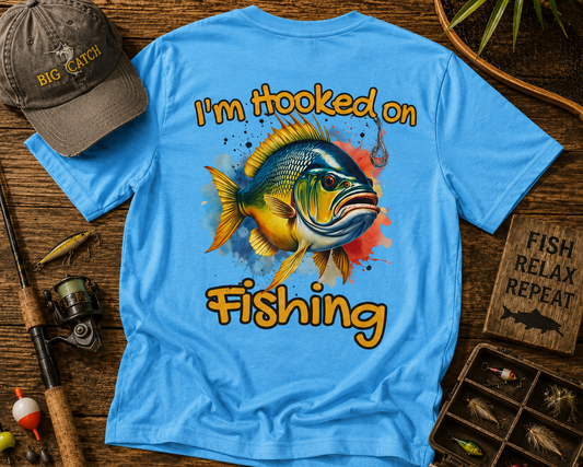 Blue t-shirt with fish graphic and 'I'm Hooked on Fishing' text, accompanied by a cap and fishing paraphernalia on a wooden surface.