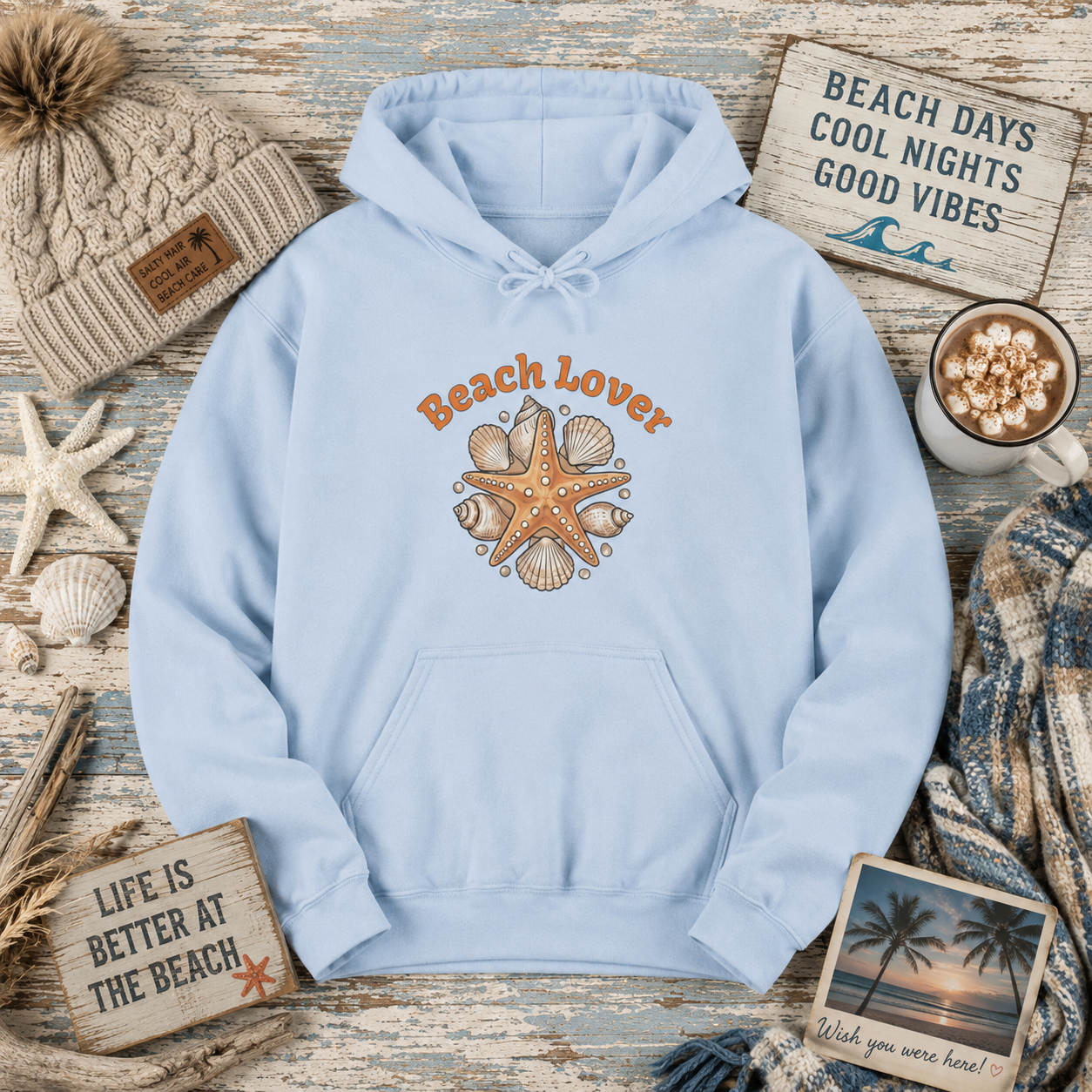 A light blue pullover hoodie with a 'Beach Lover' graphic featuring orange text and a starfish surrounded by seashells, styled in a flat lay with beach-themed props including a knit beanie, starfish, wooden signs, a mug of hot chocolate, and a scarf.
