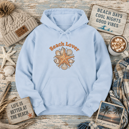 A light blue pullover hoodie with a 'Beach Lover' graphic featuring orange text and a starfish surrounded by seashells, styled in a flat lay with beach-themed props including a knit beanie, starfish, wooden signs, a mug of hot chocolate, and a scarf.