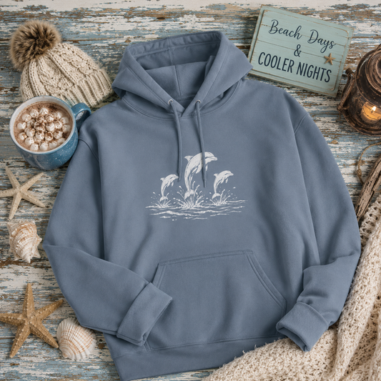 A slate blue hoodie with a white graphic of three jumping dolphins on the chest, styled in a cozy beach-themed flat lay with a knit hat, a mug of hot chocolate, starfish, seashells, and a wooden sign reading 'Beach Days & Cooler Nights'.
