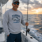 A man wearing a light gray crewneck sweatshirt with a blue 'TARPOON' fish graphic stands on a boat at sunset, holding a fishing rod. The background shows the ocean and a colorful sunset sky.