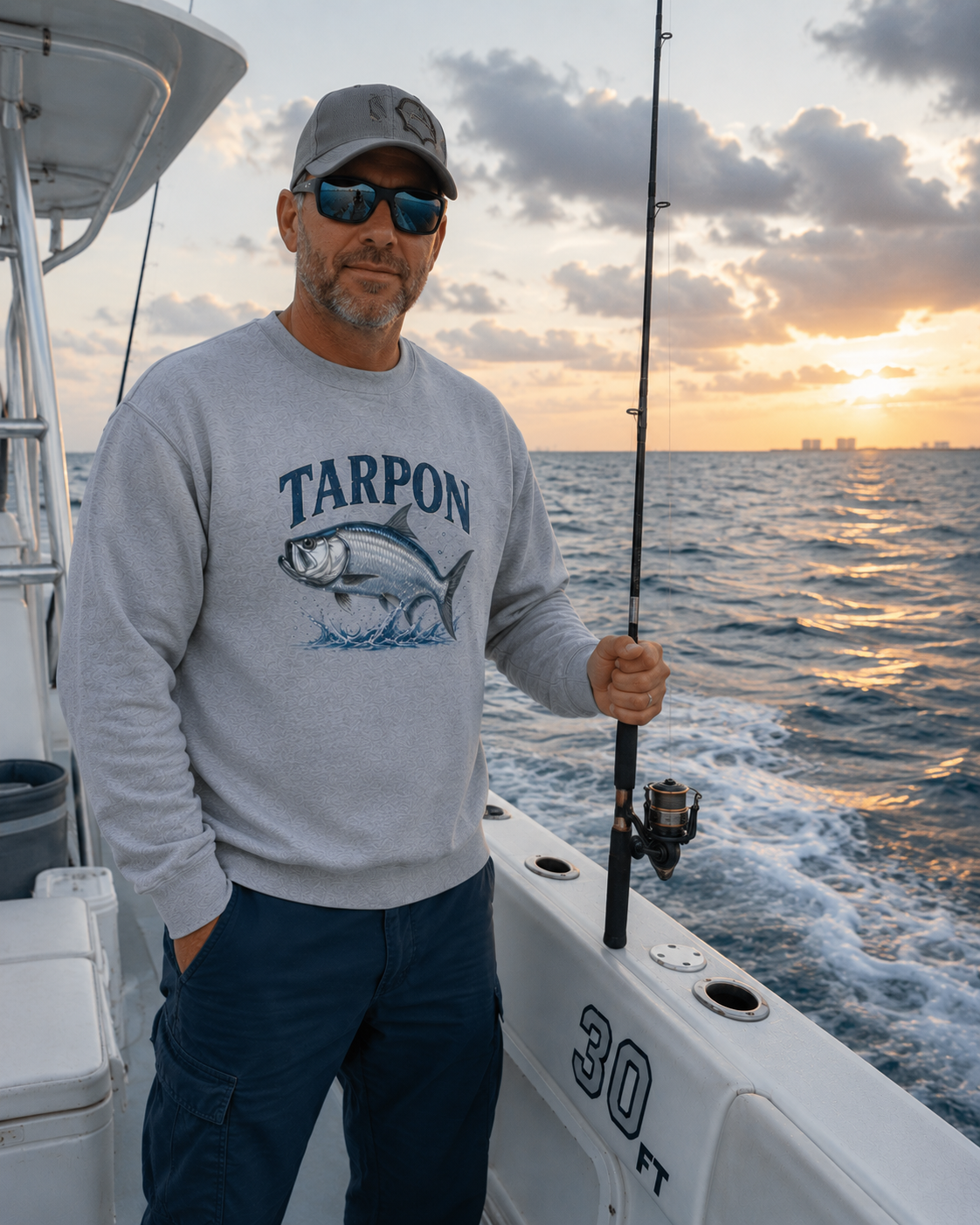 A man wearing a light gray crewneck sweatshirt with a blue 'TARPOON' fish graphic stands on a boat at sunset, holding a fishing rod. The background shows the ocean and a colorful sunset sky.
