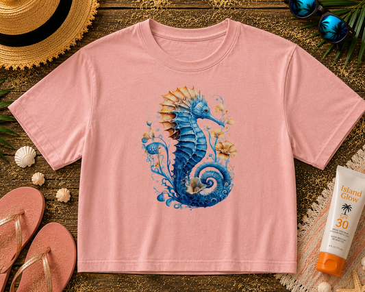 Pink t-shirt with a blue seahorse design on a wooden surface with beach accessories.