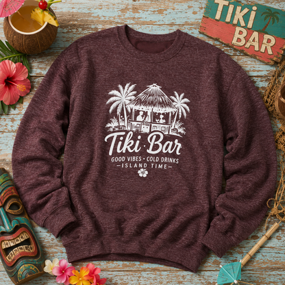 A heathered dark burgundy crewneck sweatshirt with a white graphic of a tiki bar and the text 'Tiki Bar Good Vibes - Cold Drinks - Island Time', styled in a tropical flat-lay with a maki, hibiscus flowers, and a wooden sign.