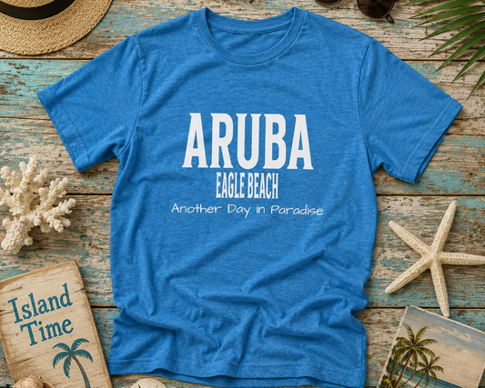 Blue t-shirt with 'ARUBA EAGLE BEACH Another Day in Paradise' text on a wooden surface with beach-themed items.