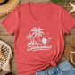 A coral red V-neck t-shirt with a white 'Bahamas' beach graphic, styled in a flat lay on a wooden surface with a straw hat, denim shorts, flip-flops, sunglasses, and seashells.