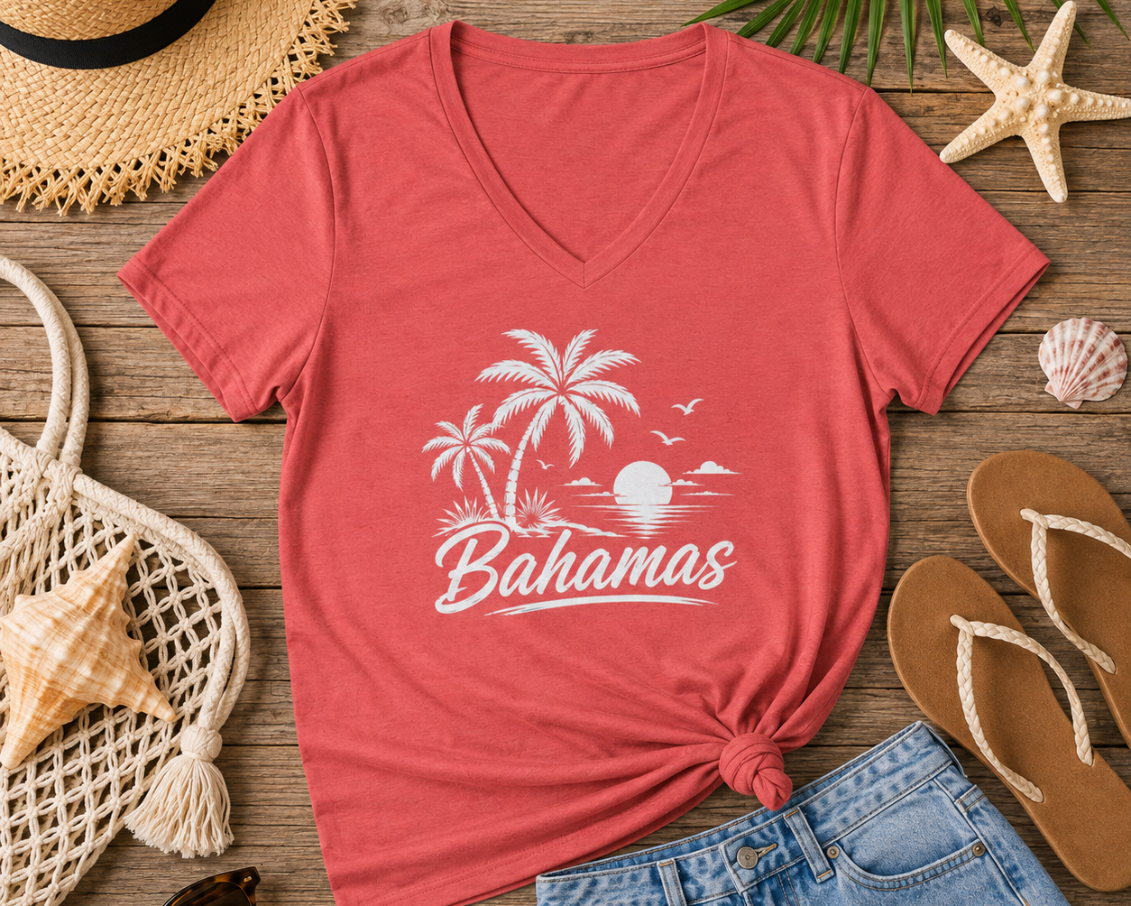 A coral red V-neck t-shirt with a white 'Bahamas' beach graphic, styled in a flat lay on a wooden surface with a straw hat, denim shorts, flip-flops, sunglasses, and seashells.