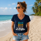 A woman standing on a sandy beach wearing a navy blue V-neck t-shirt with a colorful 'Beach Beach Beach' graphic and seashell print, paired with denim shorts.