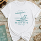 White t-shirt with 'Beach Days Best Days' design on a wooden surface with beach-themed accessories.