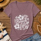 A heathered mauve V-neck t-shirt with a white 'Beach Life' floral graphic, styled in a flat lay on a wooden surface with a straw hat, sunglasses, flip-flops, denim shorts, a striped towel, and seashells.