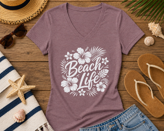 A heathered mauve V-neck t-shirt with a white 'Beach Life' floral graphic, styled in a flat lay on a wooden surface with a straw hat, sunglasses, flip-flops, denim shorts, a striped towel, and seashells.