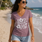 A woman walking on a sandy beach wearing a heathered mauve V-neck t-shirt with a white 'Beach Life' floral graphic, paired with denim shorts.