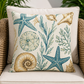 A square outdoor throw pillow with a nautical design featuring blue starfish, beige seashells, and blue-green seaweed on an off-white background, styled on a beige cushion of a wicker chair.