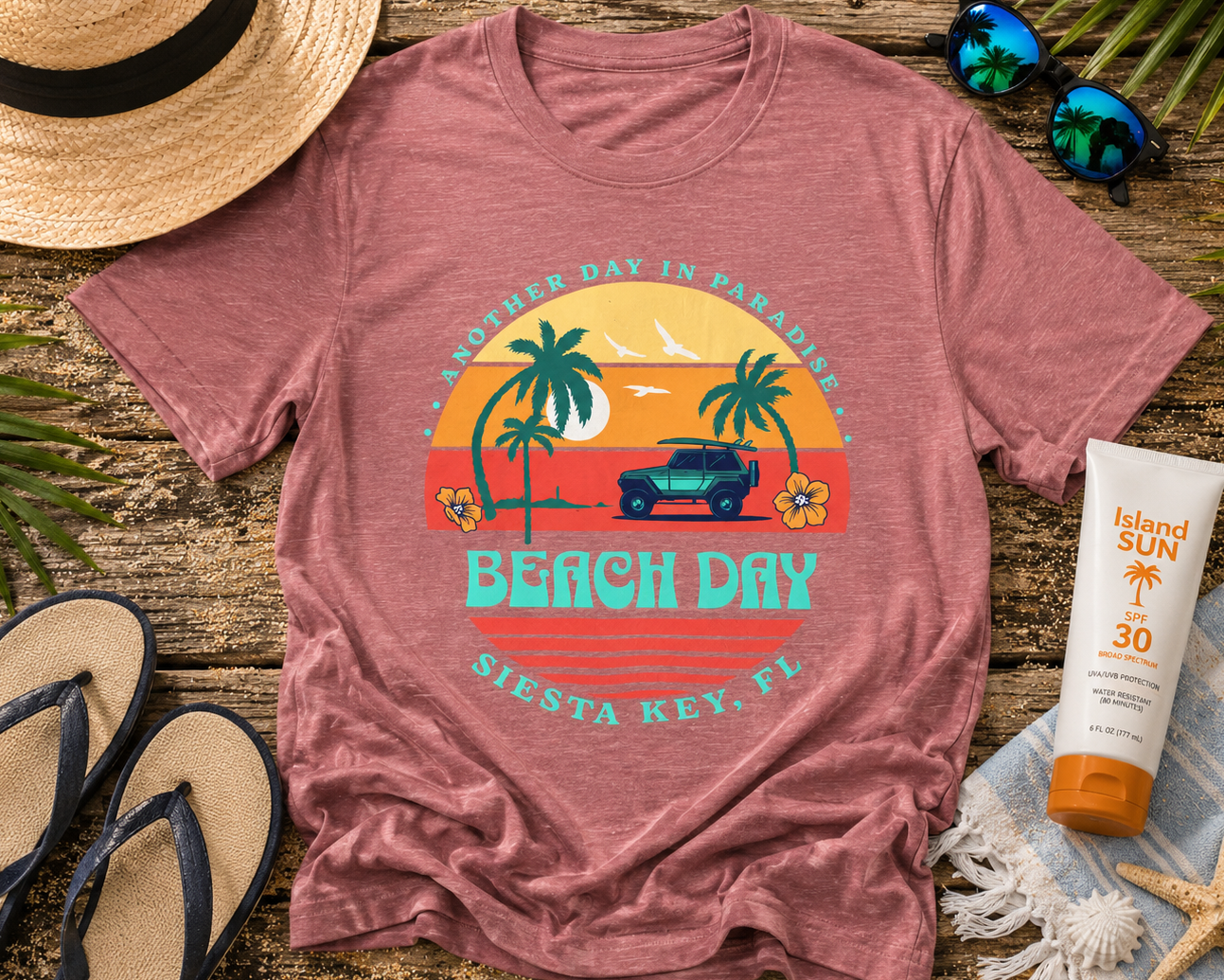 Pink t-shirt with beach-themed design on a wooden surface with sun screen, sandals, and hat.