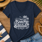 A navy blue V-neck t-shirt with a white graphic reading 'I have a Beachy Attitude' surrounded by palm trees and flowers, styled in a beach-themed flat lay with denim shorts, a straw hat, sunglasses, a striped towel, and seashells on a wooden surface.