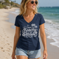 A woman walking on a sandy beach wearing a navy blue V-neck t-shirt with a white graphic that reads 'I have a Beachy Attitude' with palm trees and flowers, paired with light denim shorts.