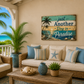 A coastal-themed living room scene with a wicker sofa and armchair, blue and striped pillows, a wooden coffee table, and a wall sign that reads 'Just Another Day in Paradise' above a beige wall. Palm trees and an ocean view are visible through the porch opening.
