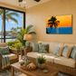 A tropical beach-themed living room featuring a canvas wall art print of a sunset over the ocean with a palm tree silhouette, hung above a beige sofa with blue and beige pillows. The room is styled with wicker furniture, plants, and a view of a canal and palm trees through large windows.