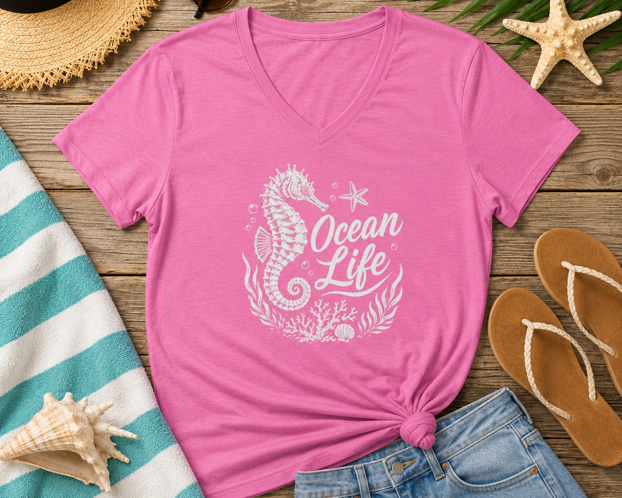 A pink V-neck t-shirt with a white 'Ocean Life' seahorse graphic, styled in a flat lay with denim shorts, a straw hat, sunglasses, flip-flops, and beach accessories on a wooden surface.