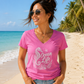 A woman wearing a bright pink V-neck t-shirt with a white seahorse and starfish graphic that reads 'Ocean Life', standing on a sunny sandy beach with palm trees and turquoise water in the background.