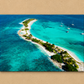 A rectangular wall art print or canvas showing an aerial view of a tropical sandbar island with turquoise water, white sand, palm trees, and a few sailboats, displayed against a beige textured wall.
