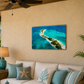 A rectangular wall art print showing a tropical island scene with turquoise water and white sand, displayed above a beige sofa in a styled living room with pillows, a lamp, and a plant.