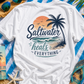 White t-shirt with 'Saltwater heals everything' design on a beach-themed background with sunglasses, flip-flops, and a tumbler.
