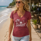 A woman wearing a heathered maroon V-neck t-shirt with a white 'Sunset' palm tree graphic, walking on a wooden boardwalk at a tropical beach.