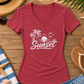 A heather red V-neck t-shirt with a white 'Sunset' beach graphic, styled in a flat lay on a wooden surface with a straw hat, sunglasses, flip-flops, denim shorts, a towel, and starfish.