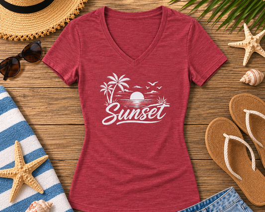 A heather red V-neck t-shirt with a white 'Sunset' beach graphic, styled in a flat lay on a wooden surface with a straw hat, sunglasses, flip-flops, denim shorts, a towel, and starfish.