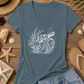 A heathered slate blue V-neck t-shirt featuring a white line art graphic of a sea turtle swimming upwards, styled in a beach-themed flat lay with denim shorts, flip-flops, a straw hat, sunglasses, and seashells on a wooden background.