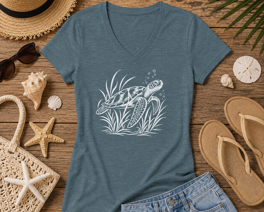 A heathered slate blue V-neck t-shirt featuring a white line art graphic of a sea turtle swimming upwards, styled in a beach-themed flat lay with denim shorts, flip-flops, a straw hat, sunglasses, and seashells on a wooden background.