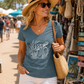 A woman wearing a slate blue V-neck t-shirt with a white sea turtle graphic, styled with white shorts, a straw hat, and sunglasses, carrying a woven straw bag on a beach market walkway.
