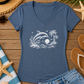 A heathered blue V-neck t-shirt with a white graphic of dolphins, palm trees, and waves, styled in a flat lay on a wooden surface with a straw hat, denim shorts, flip-flops, sunglasses, and beach accessories.