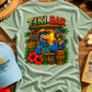 Light green t-shirt with 'Tiki Bar' design on a wooden surface with accessories.