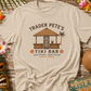 Beige t-shirt with 'Trader Pete's Tiki Bar' design on a wooden surface with tropical decorations.